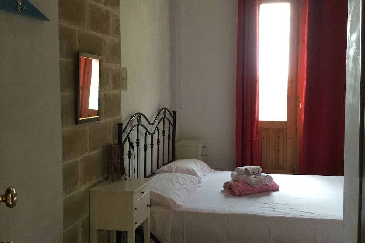A bedroom with a bed, a nightstand, a dresser, a window with red curtains, and a brick wall.