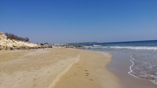 Ein weiter Sandstrand mit sanften Wellen, blauem Himmel und Felsformationen links.
