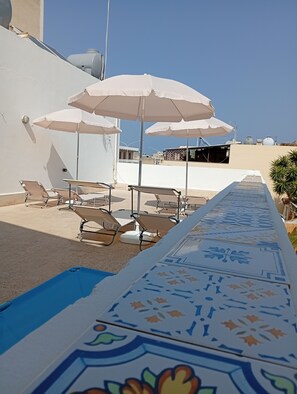Zona de piscina amb gandules i ombrel·les, un camí pavimentat i un edifici al fons.