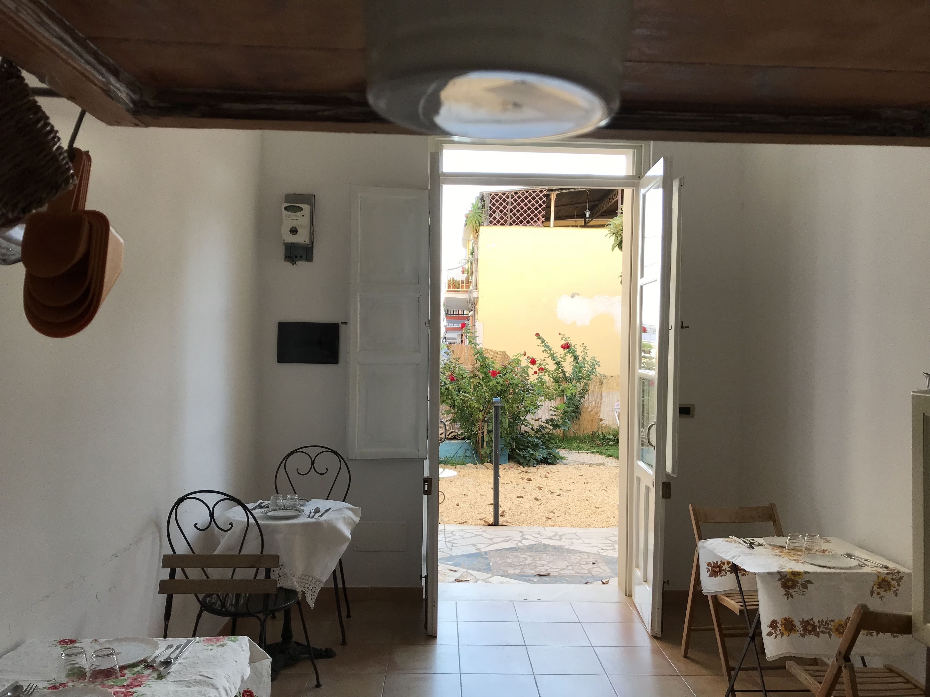 A room with a dining table set for two, a small table with a floral tablecloth, and a door leading to an outdoor area with a garden and a yellow wall.