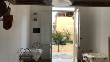 A room with a dining table set for two, a small table with a floral tablecloth, and a door leading to an outdoor area with a garden and a yellow wall.