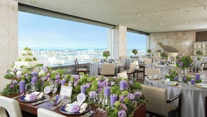 Un restaurante con techos altos y mesas dispuestas para comer, que destaca por centros de mesa con flores moradas y vistas a la ciudad.