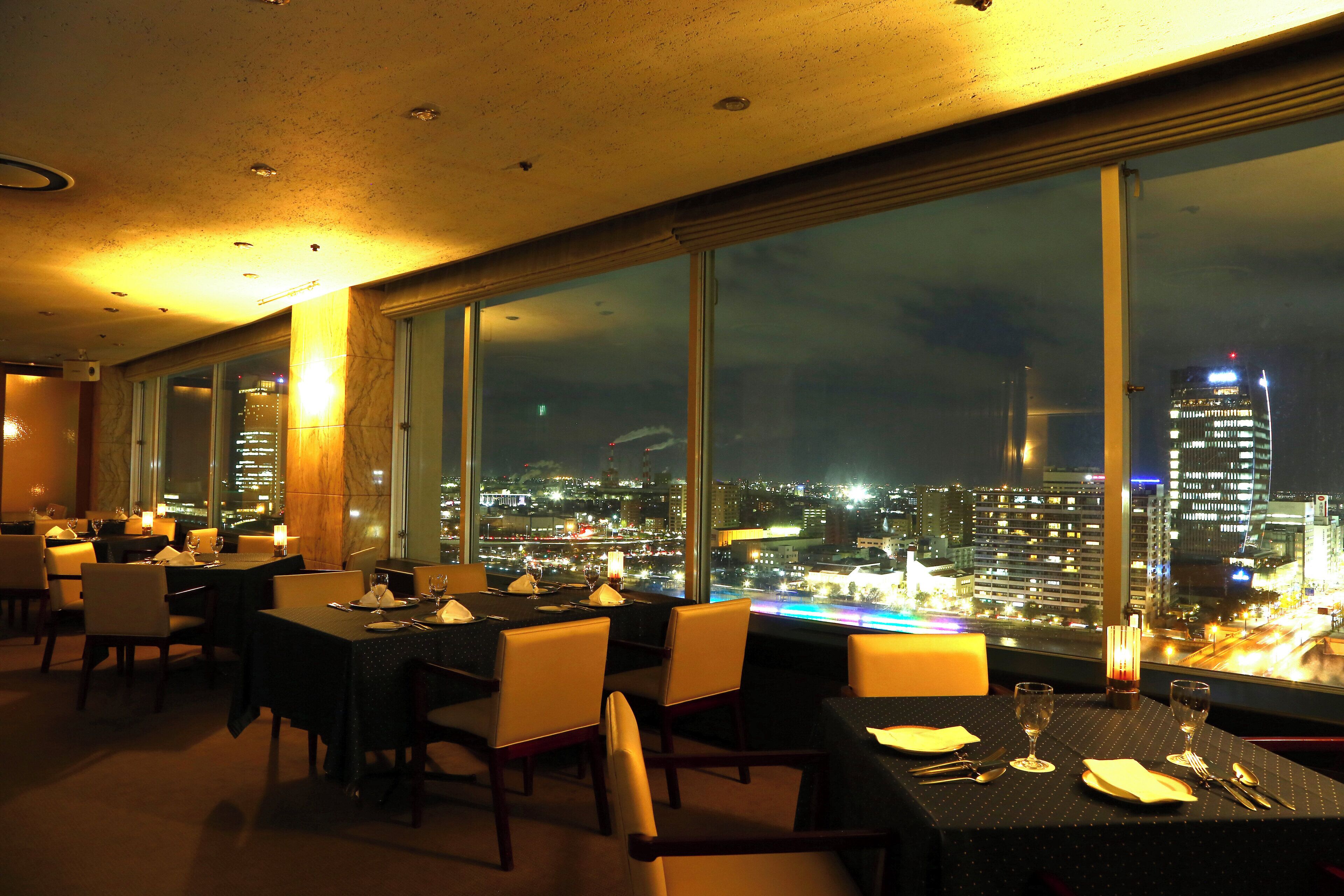Ein Restaurant mit gedeckten Tischen und großen Fenstern, die auf die beleuchtete Stadt blicken.