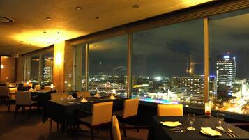 Ein Restaurant mit gedeckten Tischen und großen Fenstern, die auf die beleuchtete Stadt blicken.