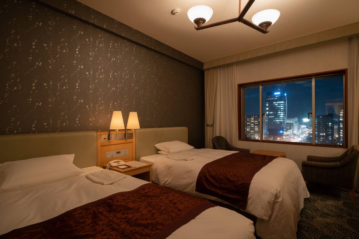 A hotel room with two beds, a nightstand with a lamp, a chair, and a window with a city view.