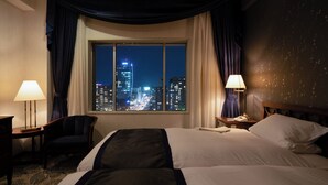 Habitación de hotel con una cama grande, dos mesitas de noche, una silla y vistas a un paisaje urbano nocturno.