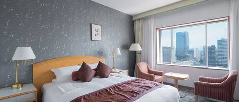 Habitación de hotel con una cama grande, dos lámparas de noche, vistas a la ciudad, una mesita pequeña y dos sillas.