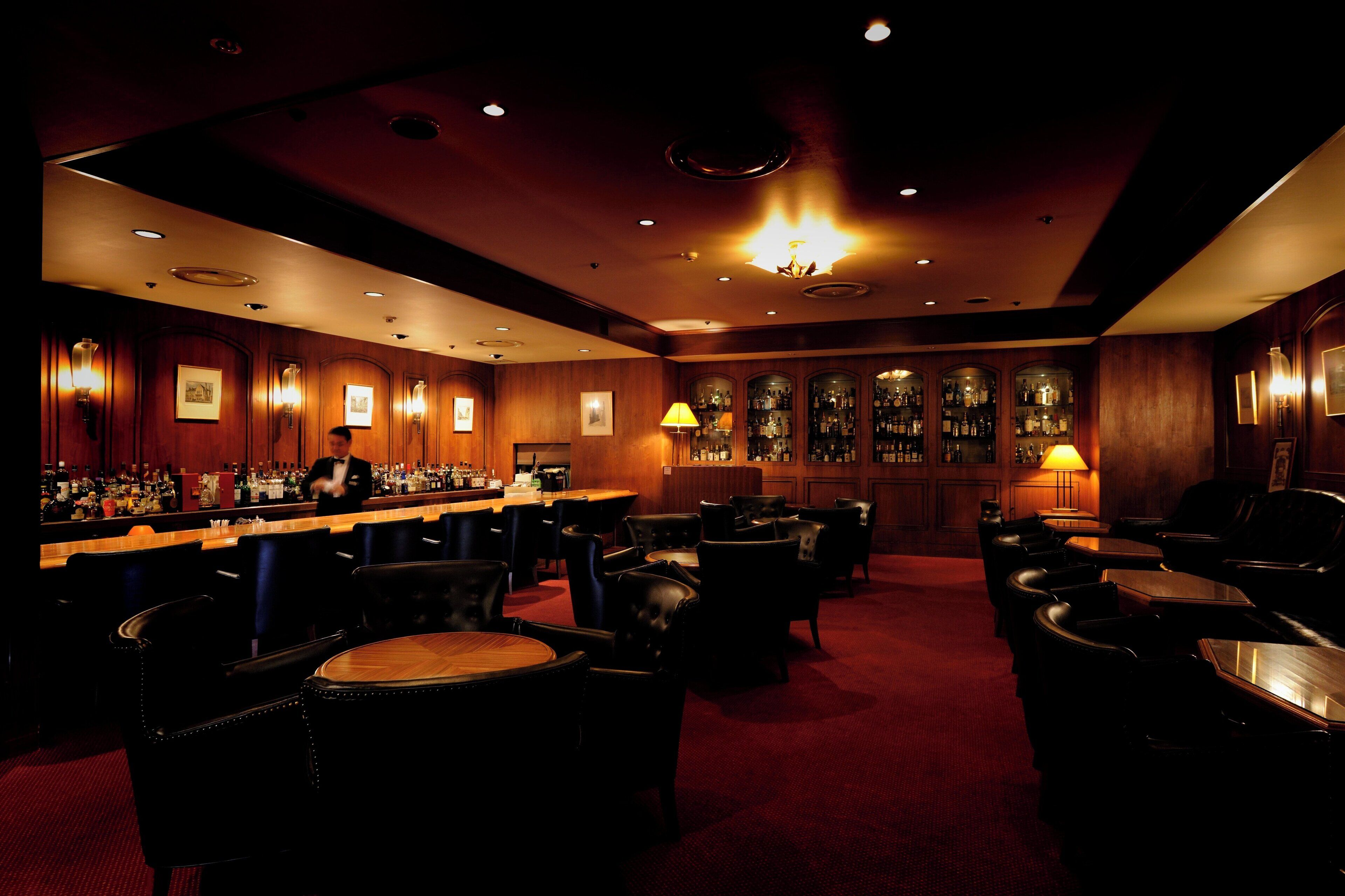 Zona de bar tenuemente iluminada, con paneles de madera, diferentes disposiciones de asientos y una barra con diversas botellas.