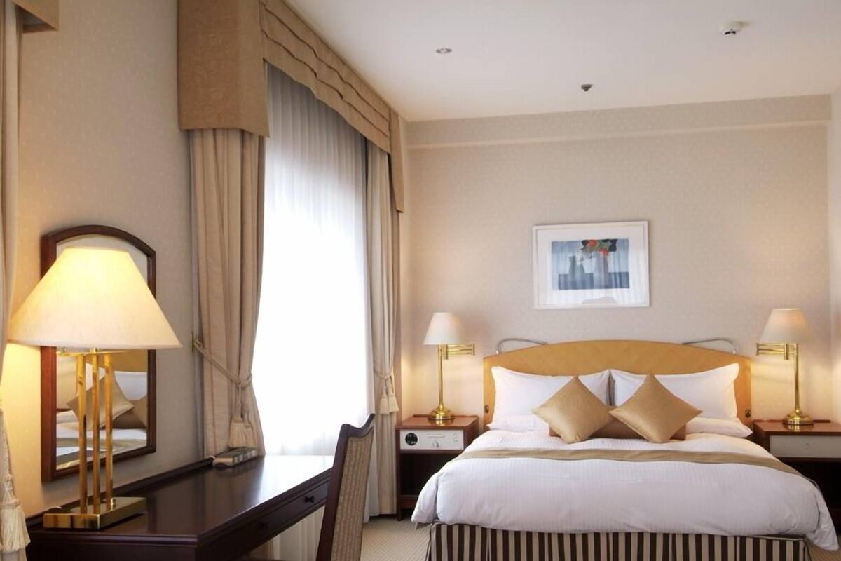 A hotel room with a bed, two bedside tables with lamps, a chair, a desk, and a large window with curtains.