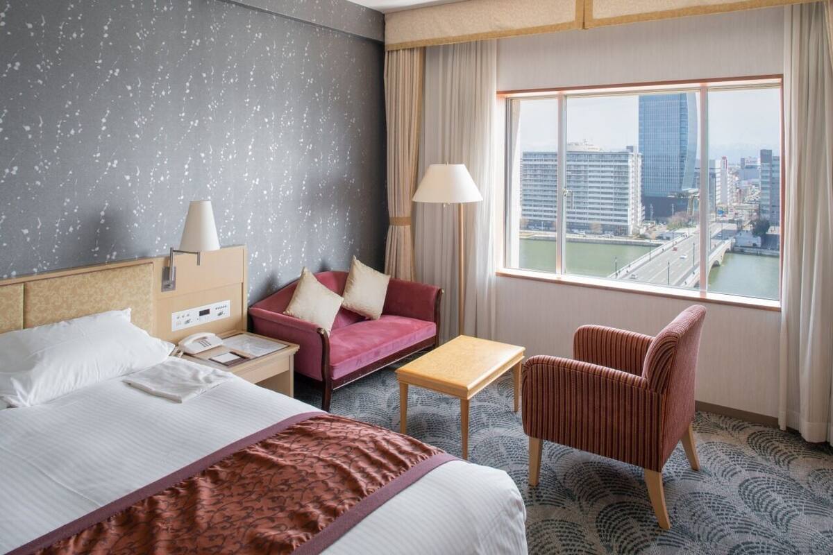 A hotel room with a bed, a desk, a chair, a sofa, and a view of a cityscape through a large window.