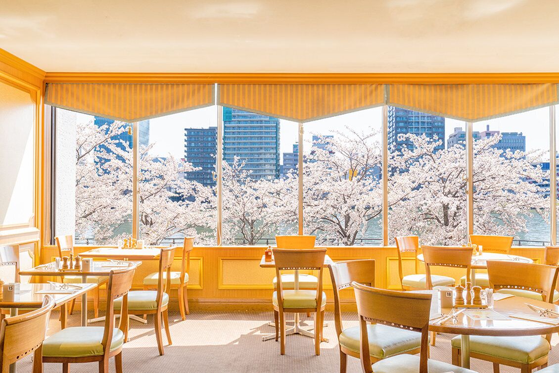 A dining area with tables and chairs, large windows with a view of cherry blossoms and buildings.