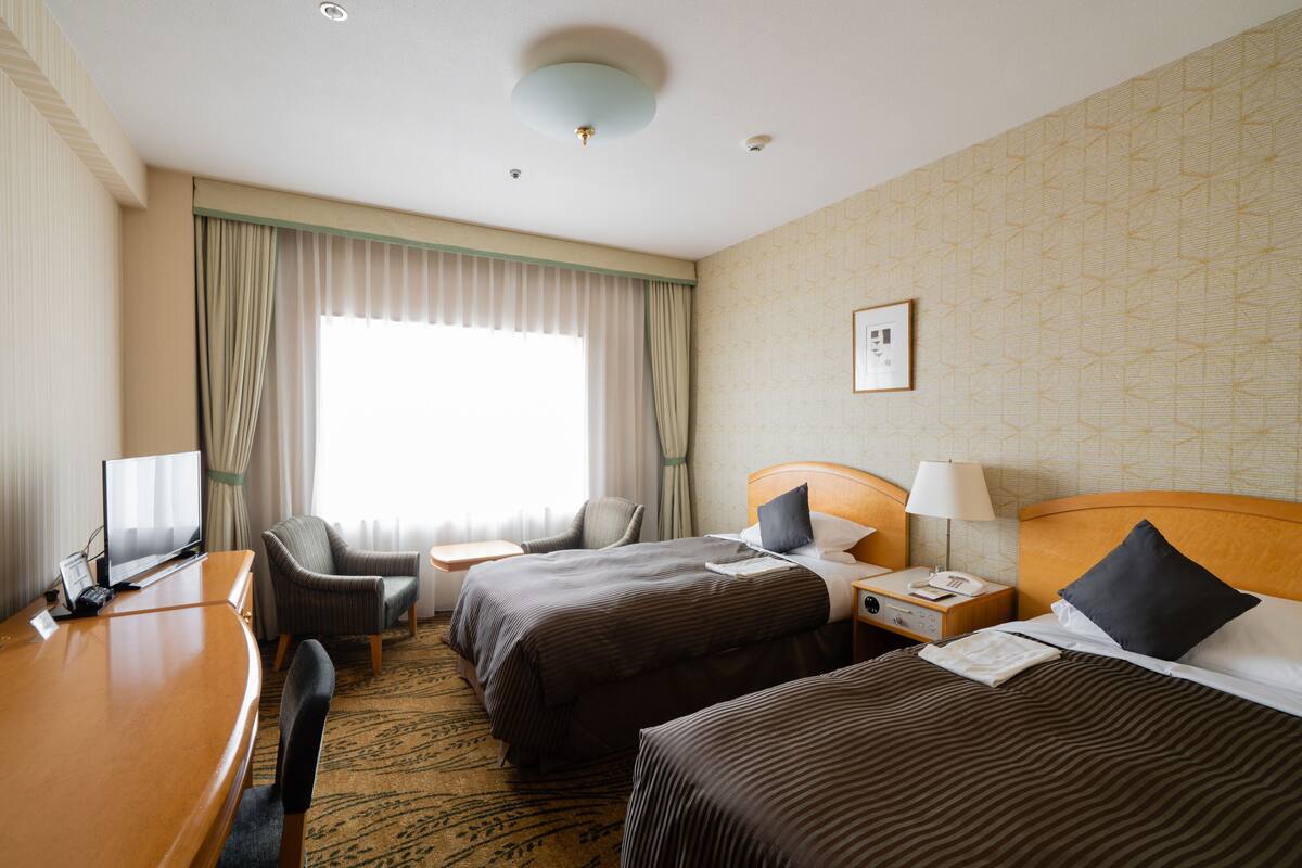 A hotel room with two beds, a desk, a chair, a desk lamp, a nightstand, and a window with curtains.