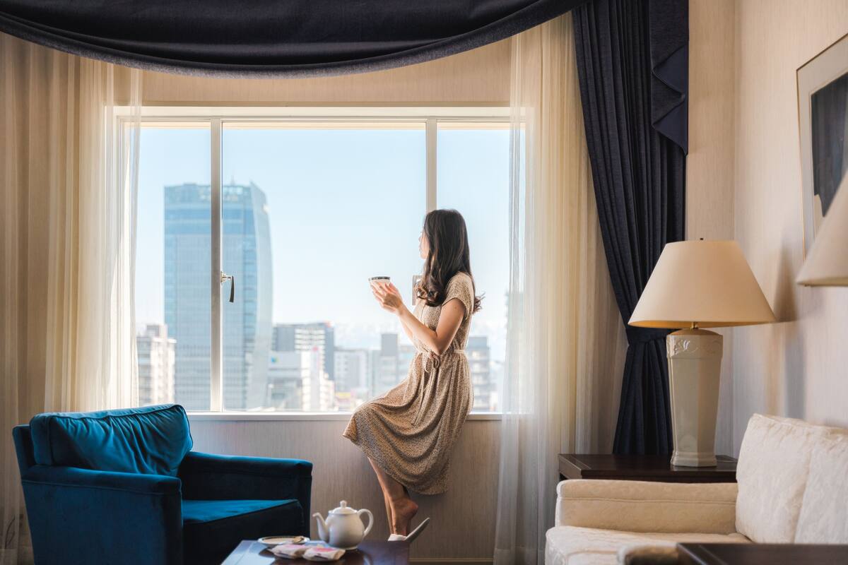 A room with a large window, a blue armchair, a white sofa, a lamp, and a tea set on a small table.