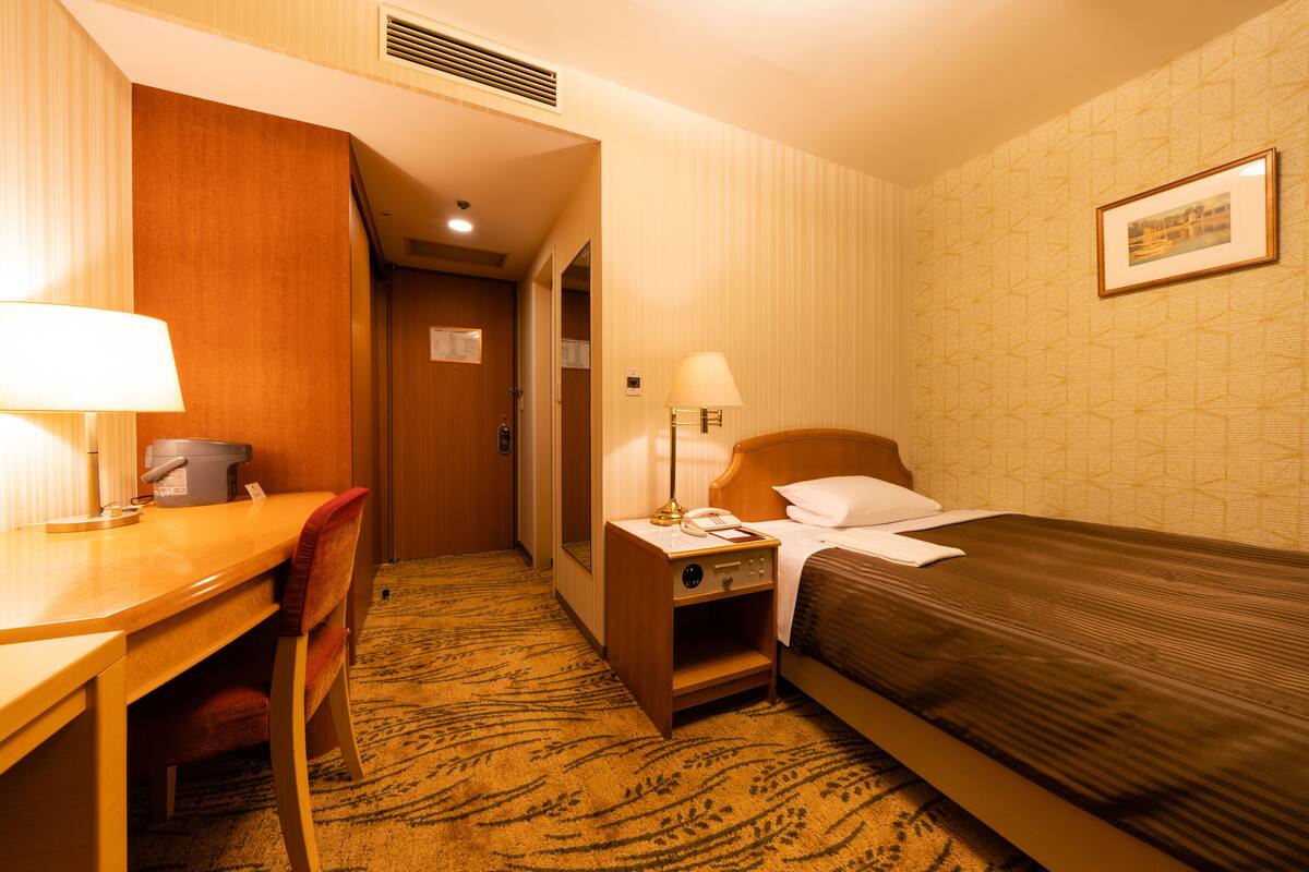 A hotel room with a bed, a desk, a chair, a nightstand with a lamp, and a framed picture on the wall.