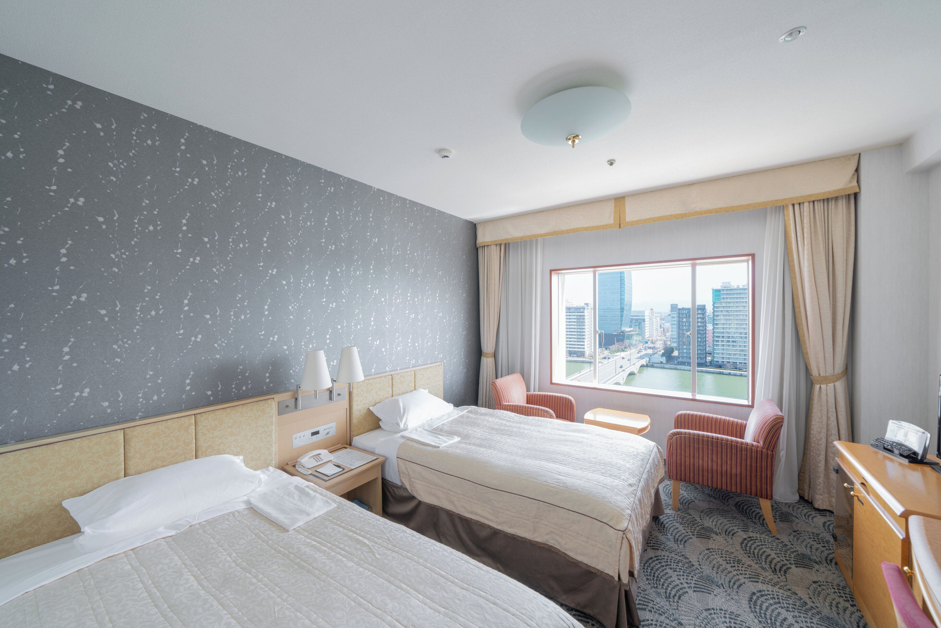 Habitación de hotel con dos camas, un escritorio, una silla, una ventana con vistas a la ciudad y una alfombra estampada.