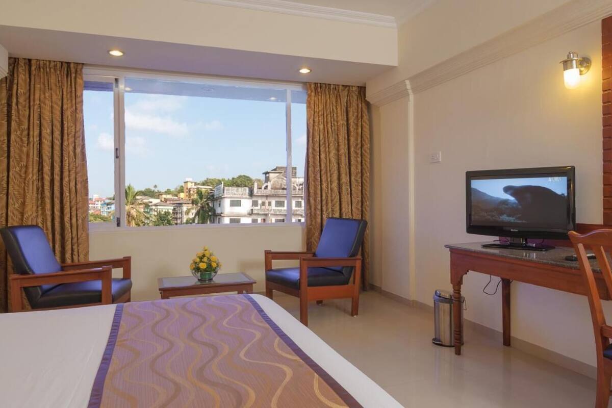 A hotel room with a bed, two armchairs, a small table with a vase, a desk with a television, and a view of buildings through a large window.