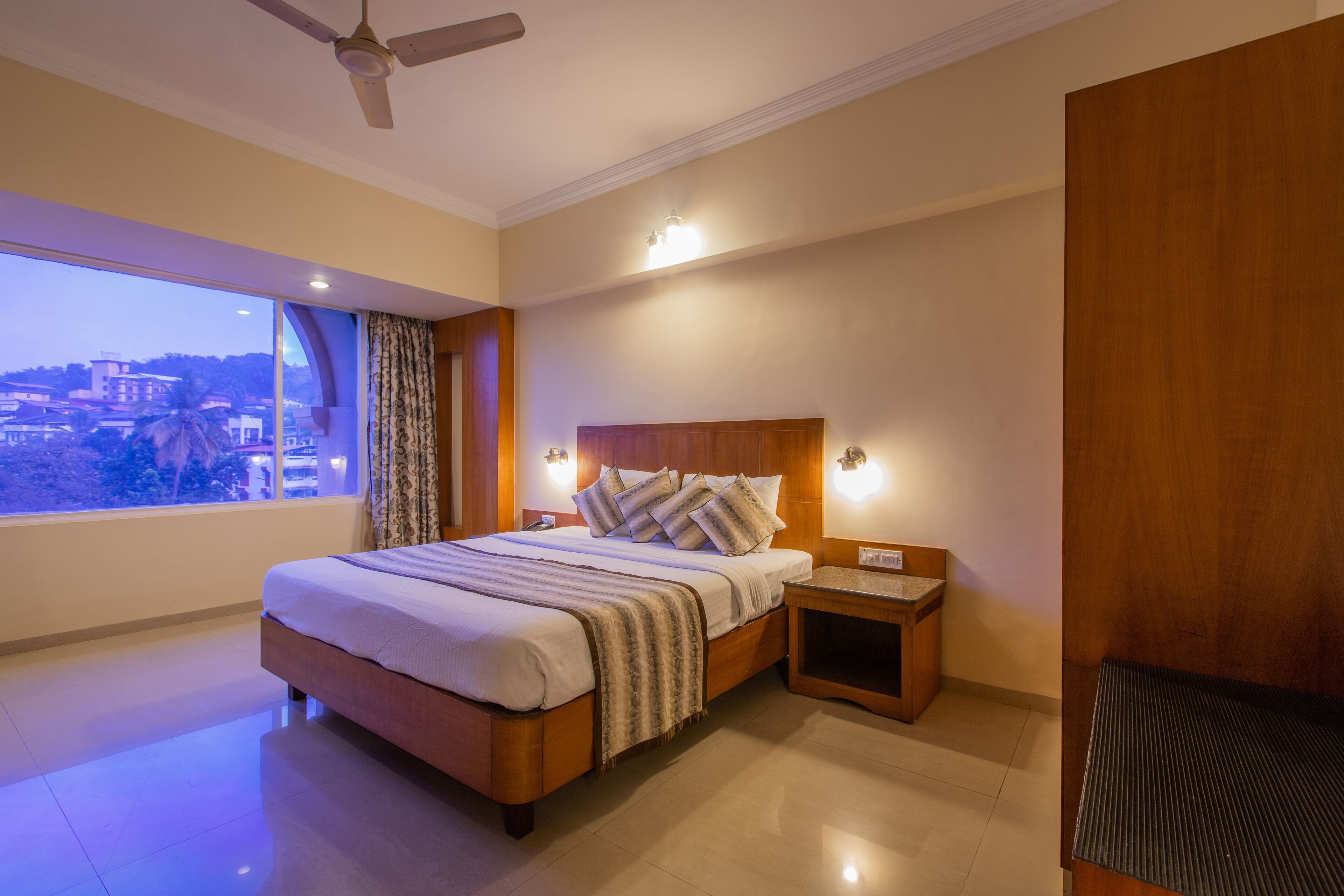 A hotel room with a large bed, bedside tables, a ceiling fan, and a view of a cityscape through a large window.