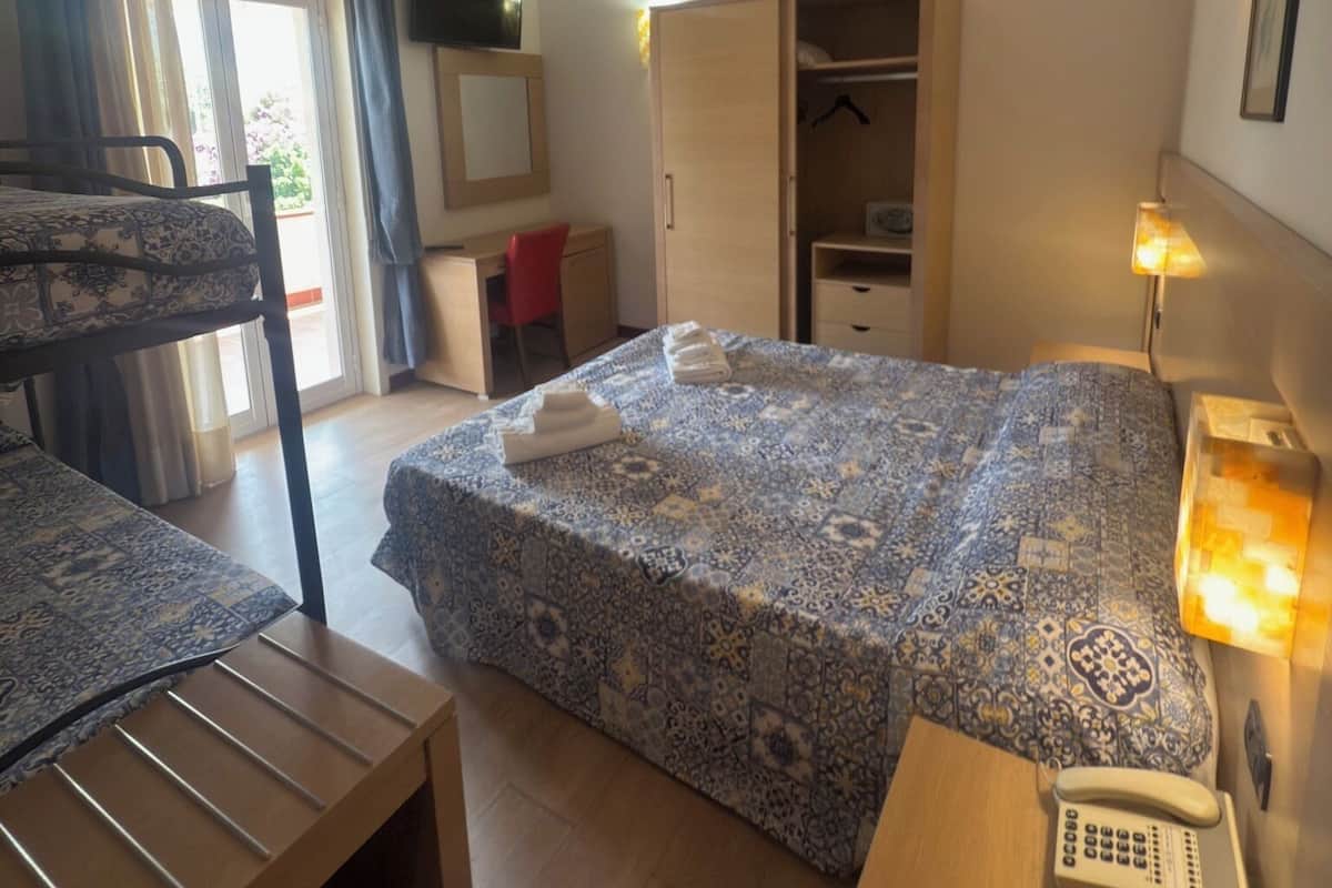 A hotel room with a bed, a desk, a chair, a wardrobe, and a bedside table with a lamp and a phone.
