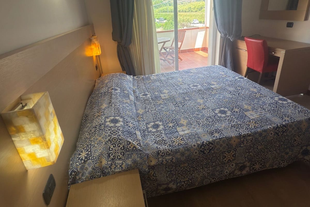 A hotel room with a bed, a desk with a telephone, a chair, a desk lamp, and a view of the outdoors through a sliding door.