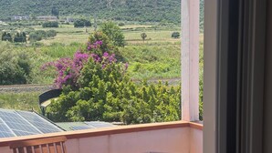 Uma varanda com mesa e cadeira, com vista para uma paisagem montanhosa com vegetação e flores roxas.