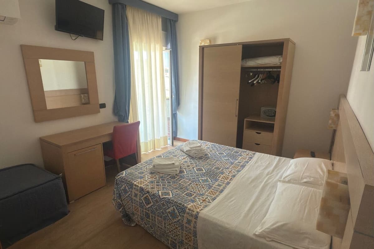 A hotel room with a bed, a desk, a wardrobe, a mirror, a TV, and a telephone.