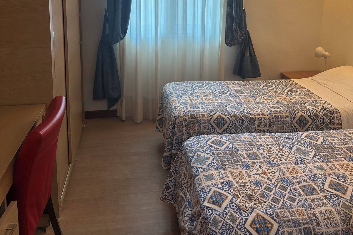 A hotel room with two beds, a desk, a chair, a wardrobe, and a window with curtains.