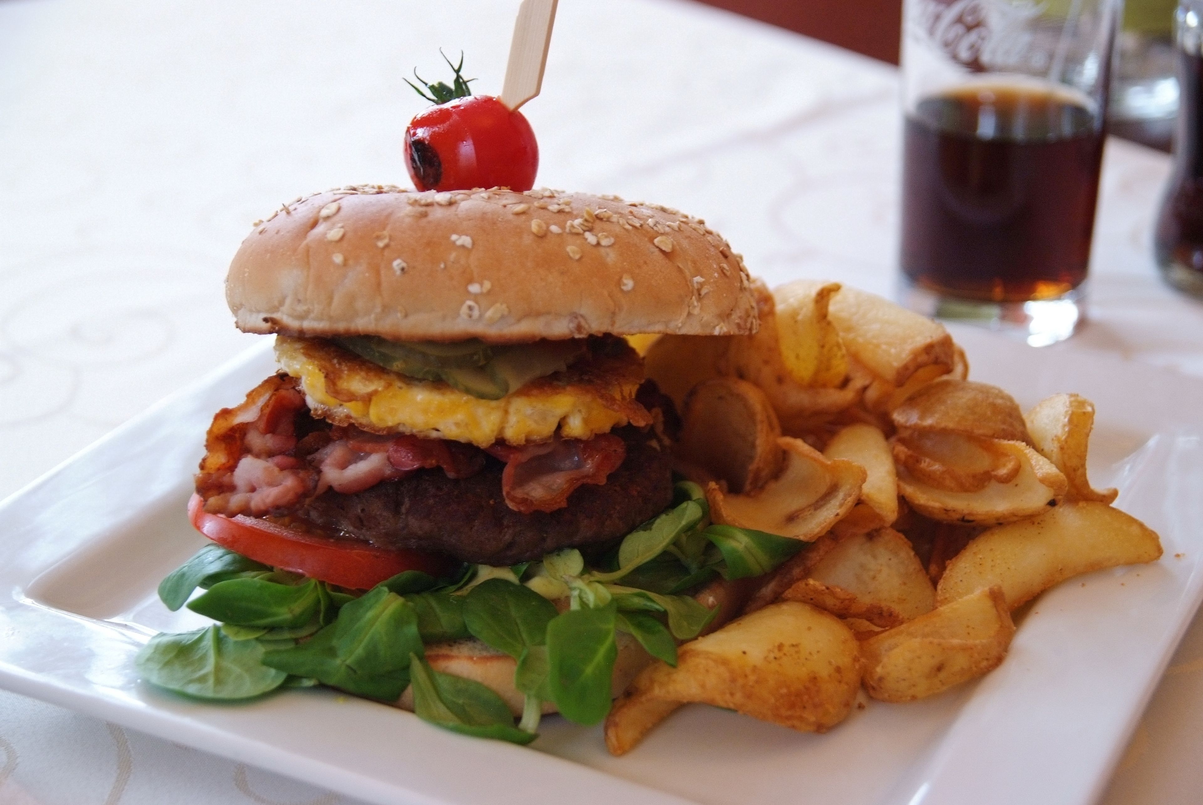A burger with a sesame seed bun, bacon, lettuce, tomato, and cheese, accompanied by a side of potato wedges and a glass of dark beverage.