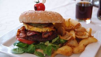 A burger with a sesame seed bun, bacon, lettuce, tomato, and cheese, accompanied by a side of potato wedges and a glass of dark beverage.