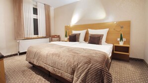 Una habitación de hotel con una cama grande, cabecera de madera, dos mesitas de noche con lámparas, una ventana con cortinas y un radiador.