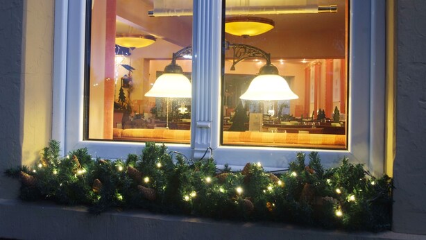 Una ventana que muestra un interior cálidamente iluminado, decorado con luces navideñas y piñas.
