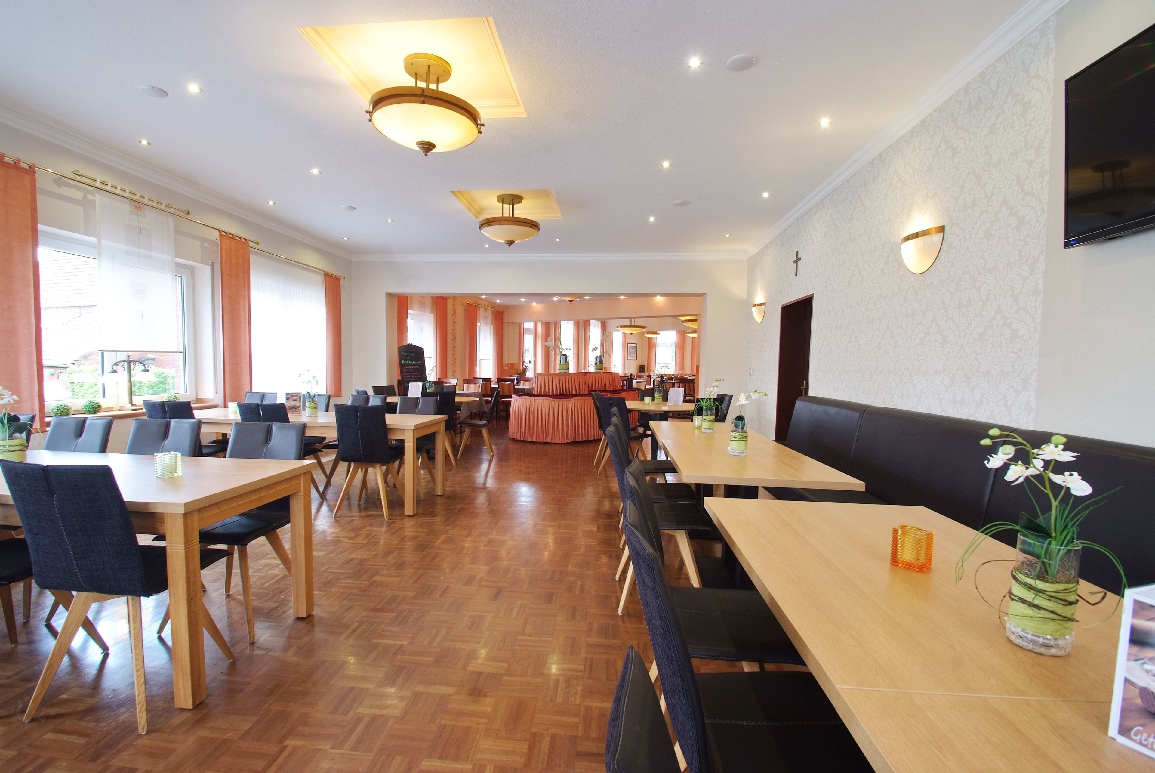 A spacious dining area with wooden tables and chairs, a central seating area with a pink tablecloth, and a cross on the wall.