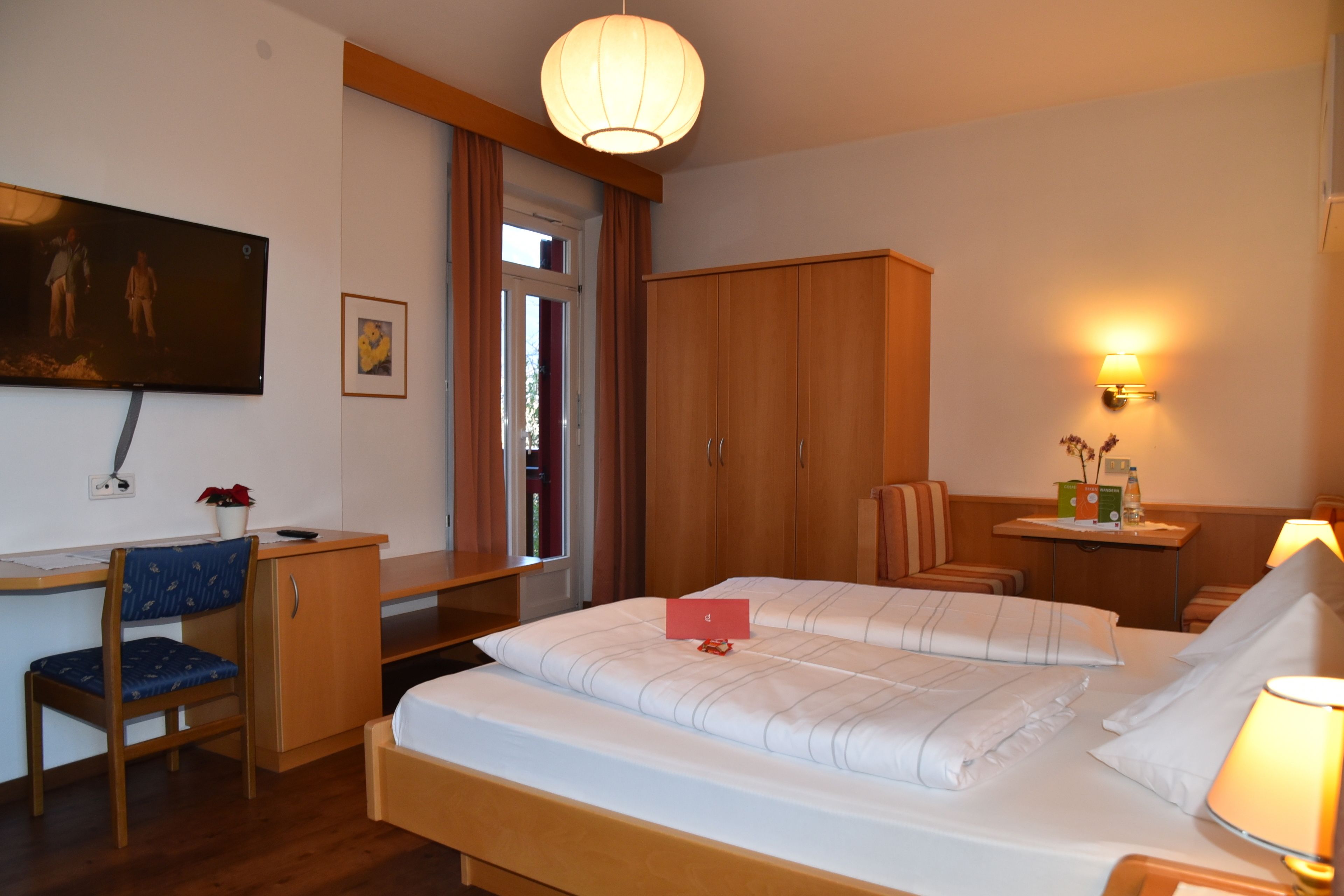 A hotel room with a bed, desk, chair, wardrobe, and a TV on the wall.