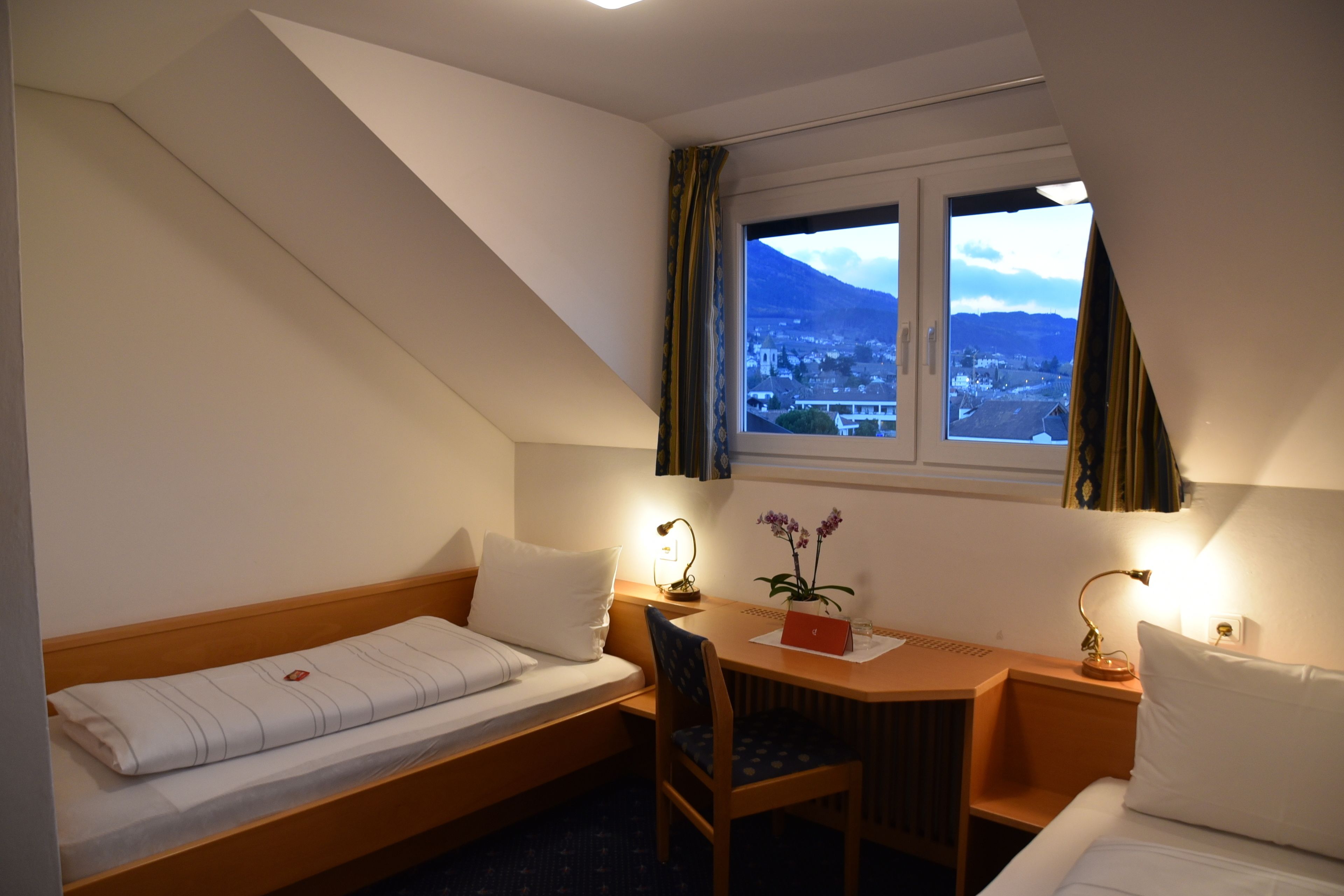 A hotel room with a bed, a desk, a chair, a window with curtains, and a view of a mountain and town.
