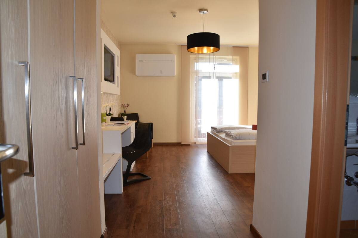 Une chambre d’hôtel moderne avec un plancher en bois, un lit, un bureau avec une chaise, un luminaire suspendu et une fenêtre laissant entrer la lumière naturelle.