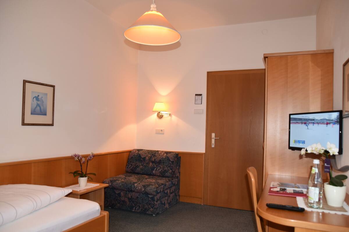 Une chambre d’hôtel avec un lit, un fauteuil à motifs, un bureau avec un ordinateur, une lampe et une petite table avec une télécommande.