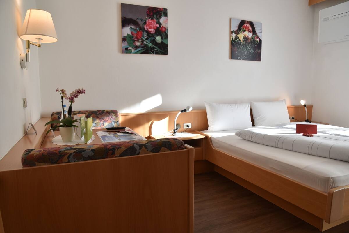 Une chambre d’hôtel moderne avec un lit en bois, une table de chevet avec une lampe, un petit bureau et une chaise. On y trouve deux lampes fixées au mur, une plante en pot et deux tableaux floraux encadrés.