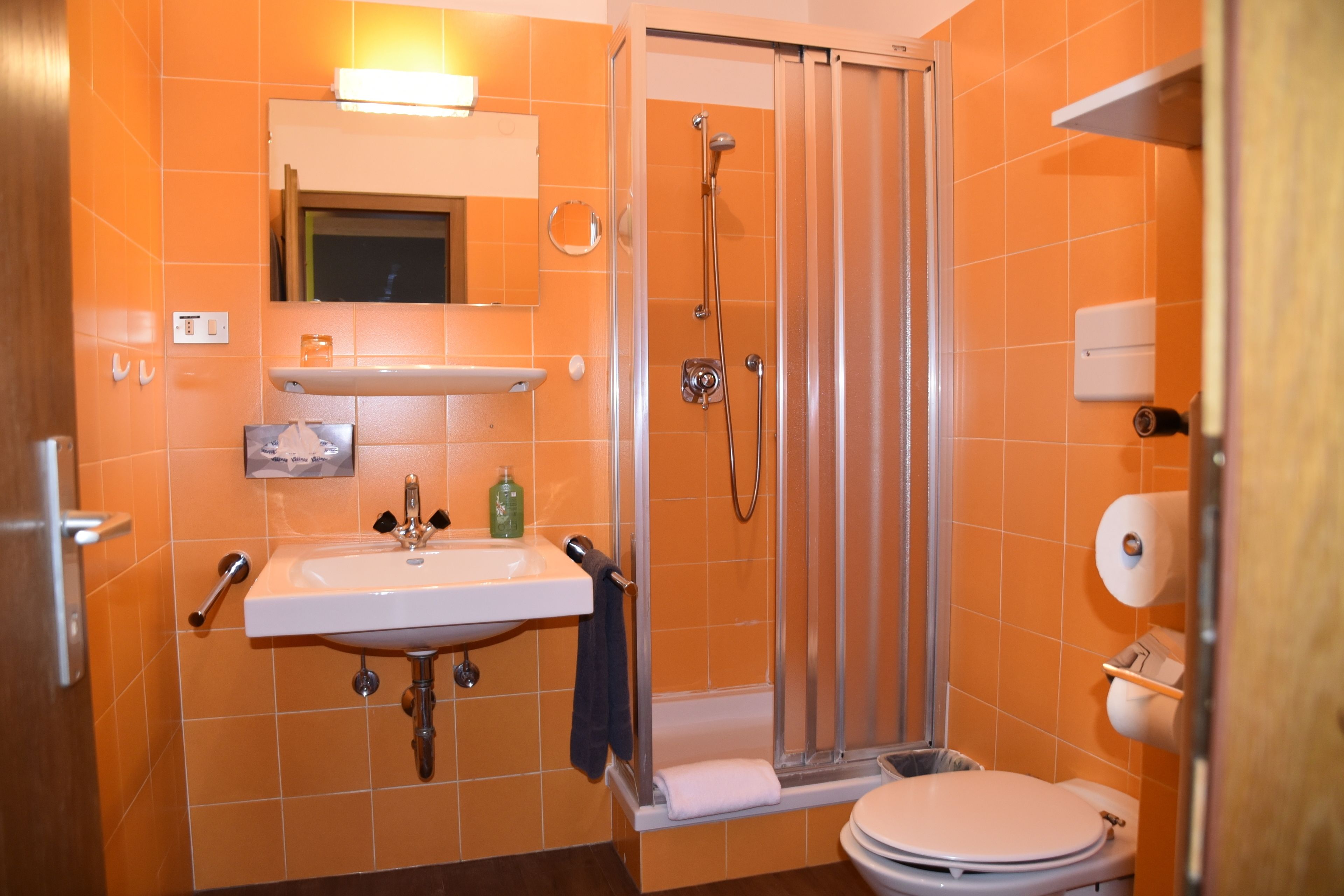 Un baño con pared de azulejos anaranjados, un lavabo blanco, un espejo, una cabina de ducha y un inodoro.