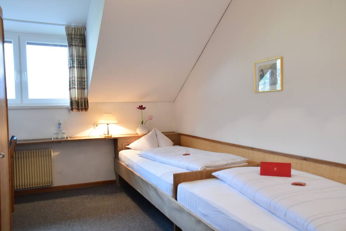 Une petite pièce bien éclairée, avec deux lits simples, une table de chevet avec une lampe, un radiateur et une fenêtre avec des rideaux.