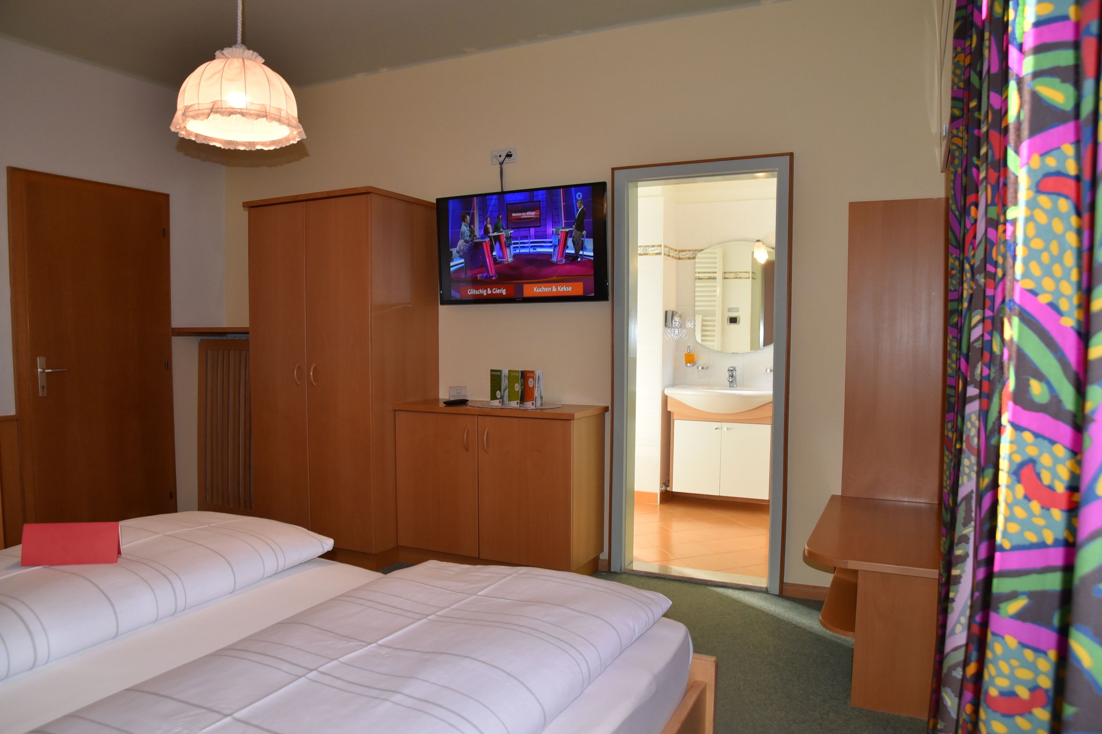 A hotel room with a bed, wooden wardrobe, a television displaying a show, and a bathroom visible through an open door.