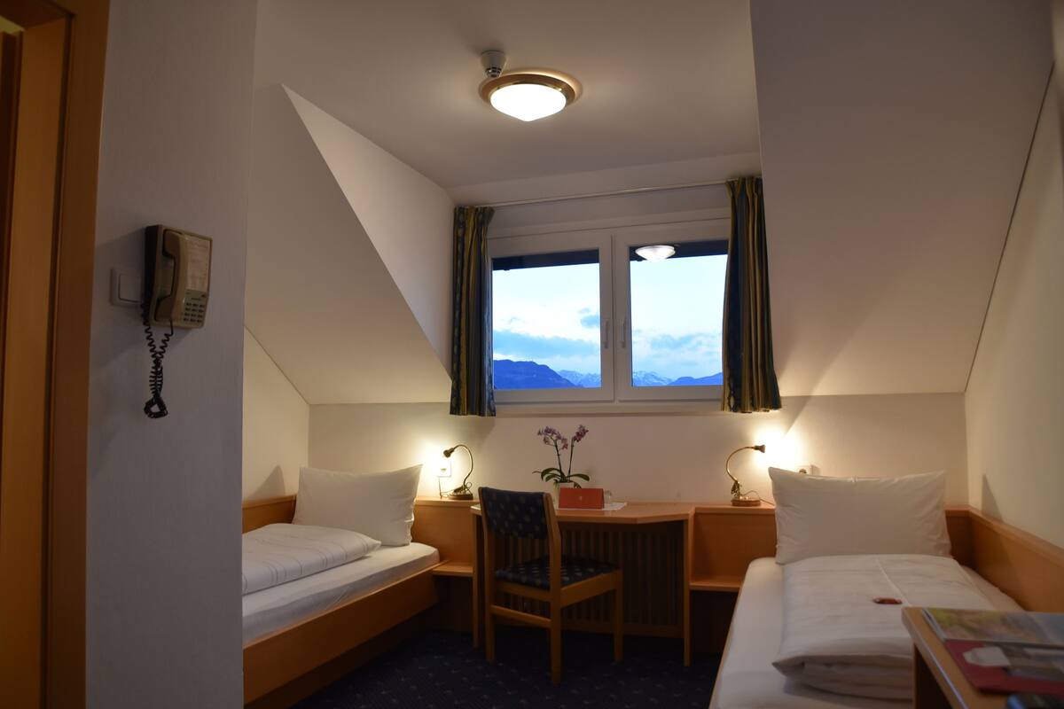 Une chambre d’hôtel comprenant un lit, un bureau avec une chaise, un téléphone et une fenêtre avec des rideaux.