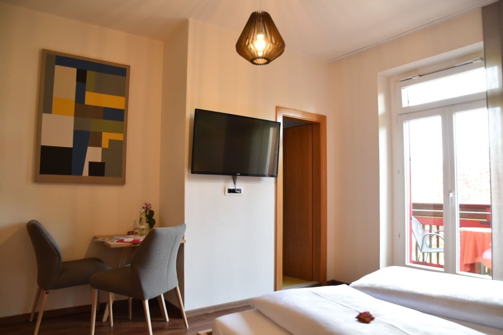 A hotel room with a bed, a desk with a chair, a TV mounted on the wall, and a balcony with a table and chairs.