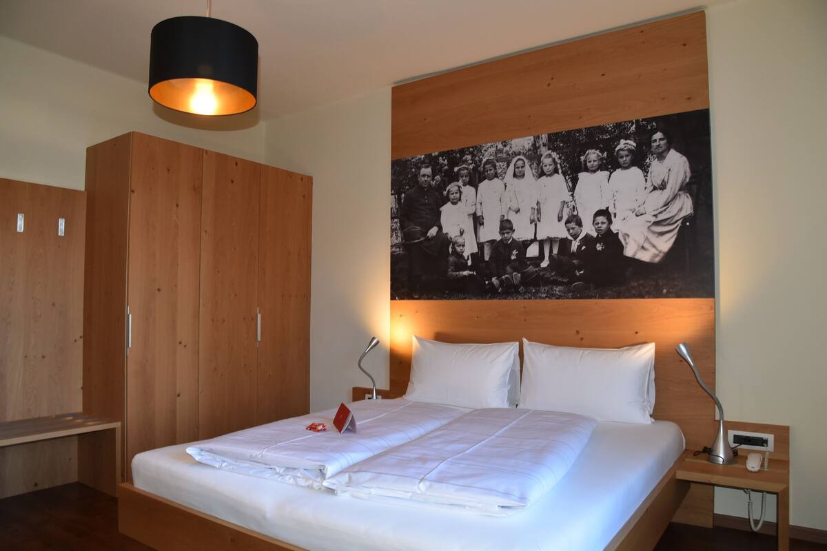 Un lit bien fait, avec du linge de lit blanc, une tête de lit en bois, une lampe suspendue noire fixée au mur, une table de chevet avec une lampe, et une photo de famille en noir et blanc encadrée au-dessus du lit.