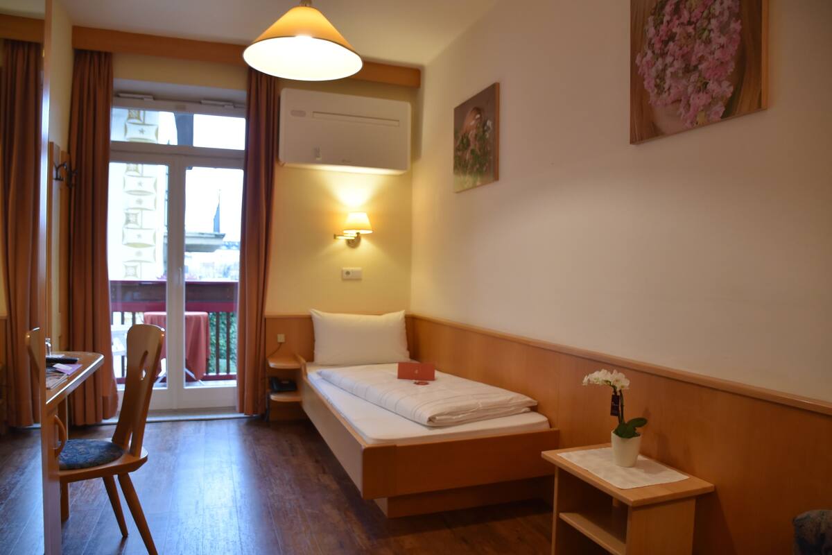 Une chambre d’hôtel comprenant un lit, un bureau, une chaise, une table de chevet, une lampe, une plante en pot et une fenêtre avec des rideaux.