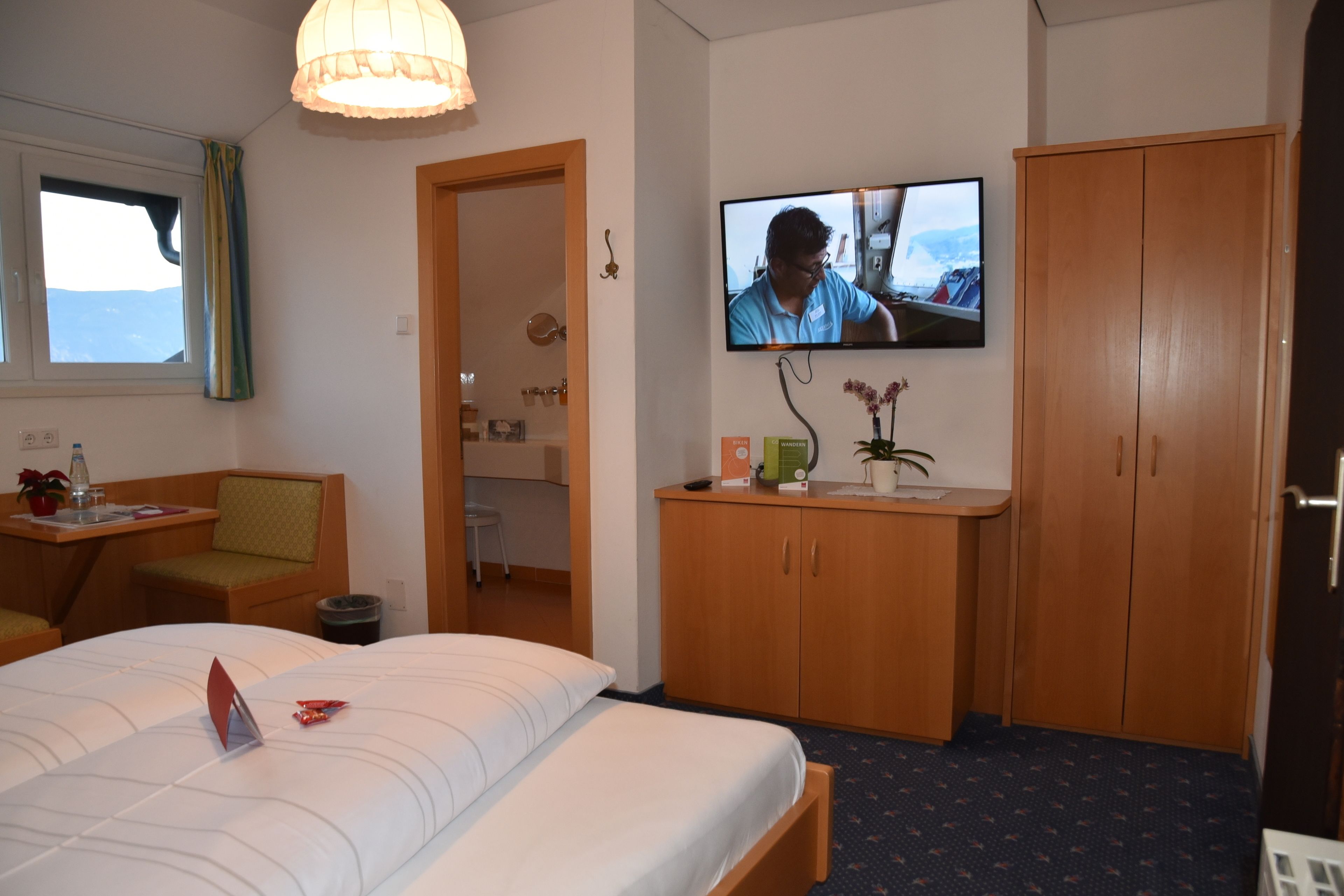 A hotel room with a bed, a desk with a chair, a television mounted on the wall, and a wardrobe.