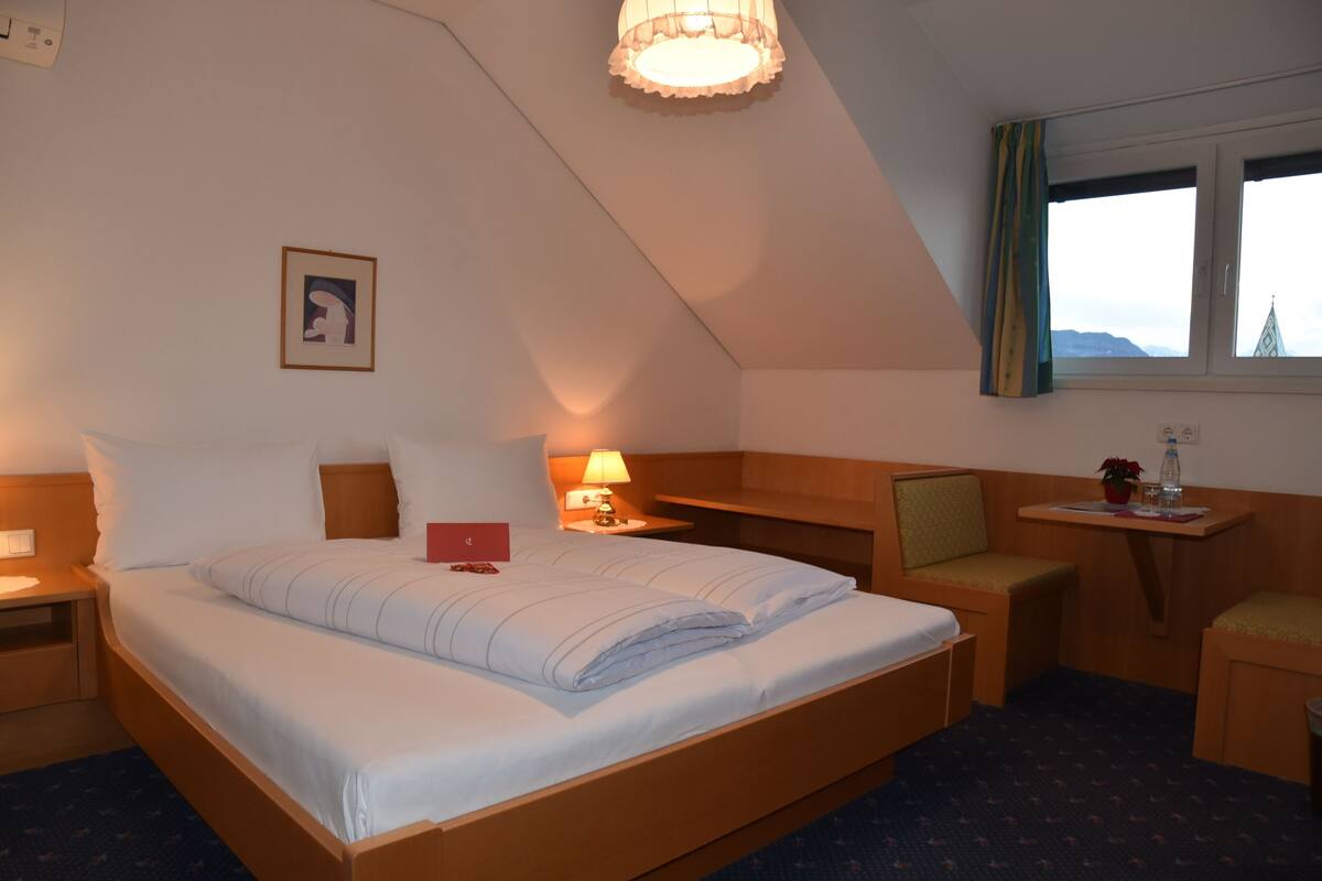 Une chambre d’hôtel avec un grand lit, un bureau, une chaise, une fenêtre avec des rideaux et une petite table sur laquelle se trouvent une plante et une bouteille.