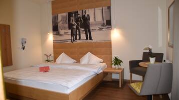 Una habitación de hotel moderna con una cama de madera, una mesita de noche pequeña con una planta, una mesa redonda con dos sillas y una fotografía enmarcada en la pared.