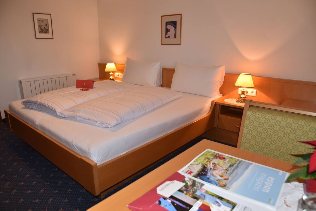 Une chambre d’hôtel avec un lit, deux tables de chevet avec des lampes, une petite table avec un magazine et un radiateur.