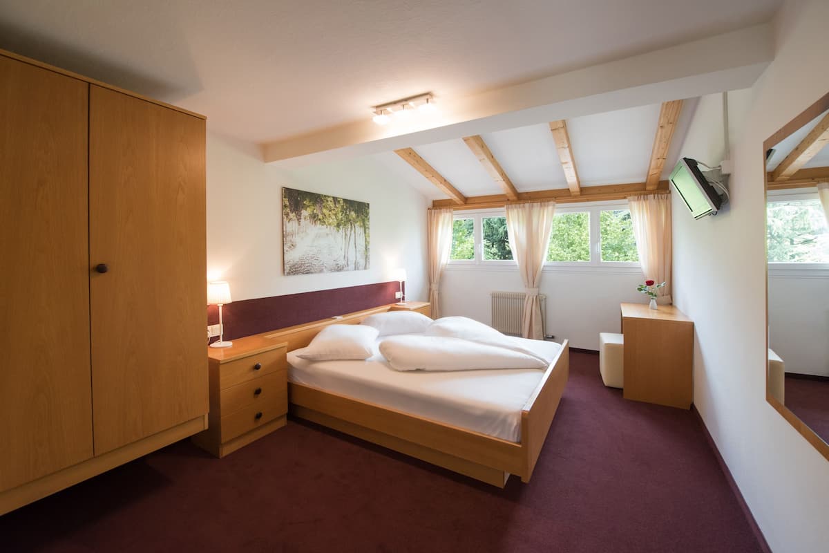 A bedroom with a wooden bed, a nightstand, a bedside table with a lamp, a painting on the wall, a window with curtains, and a television mounted on the wall.