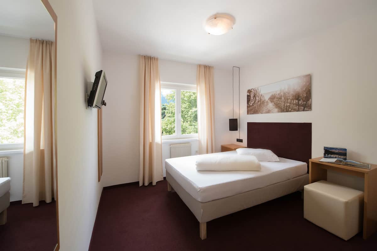A hotel room with a bed, a bedside table, a nightstand, a window with curtains, a radiator, and a wall-mounted artwork.