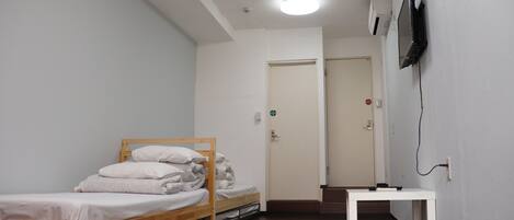 Una habitación pequeña y limpia con una cama individual, una mesa blanca y un televisor montado en la pared.